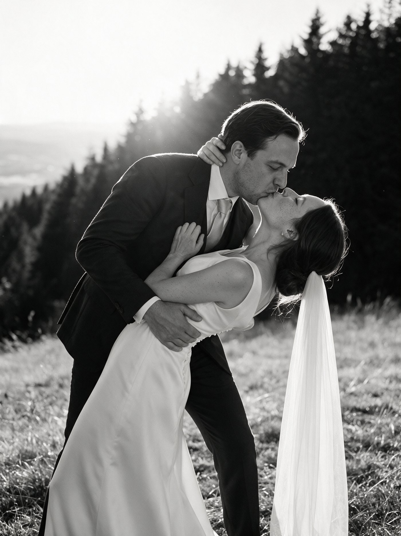 A groom dips his bride as they kiss in a mountain meadow, backlit by low evening sun with pine forest behind them