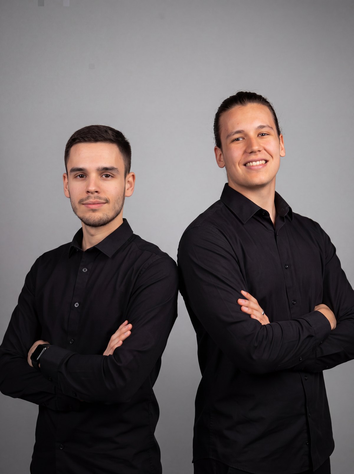 Lan and Jure of Flanner Wedding Films, standing back to back in black shirts against a grey studio backdrop